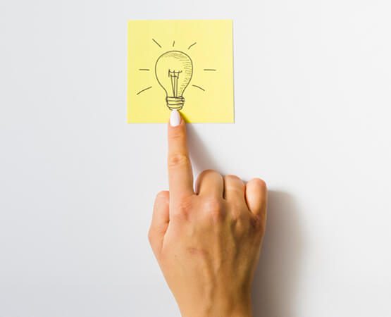 A hand sticking a yellow note with a drawn lightbulb icon onto a clear surface, representing a moment of creative insight, a breakthrough idea, or the strategic solution delivered.